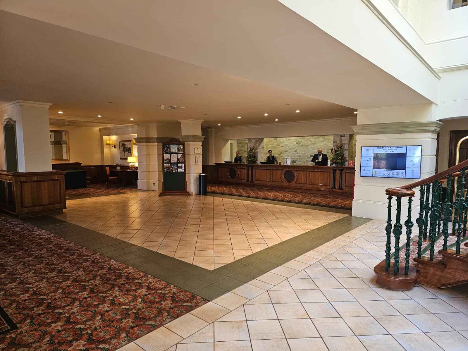 Hotel de France reception today — patterned carpet, wooden desk panelling, and a digital welcome screen.